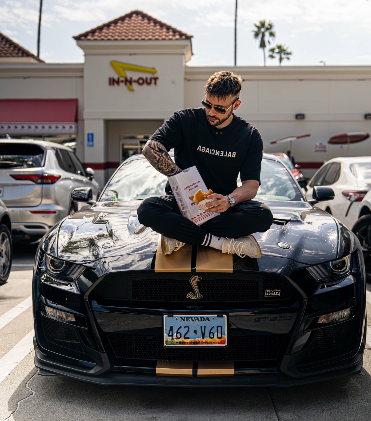 Philipe Reis sitting on the bonnet of a black sports car outside an In-N-Out, eating a burger