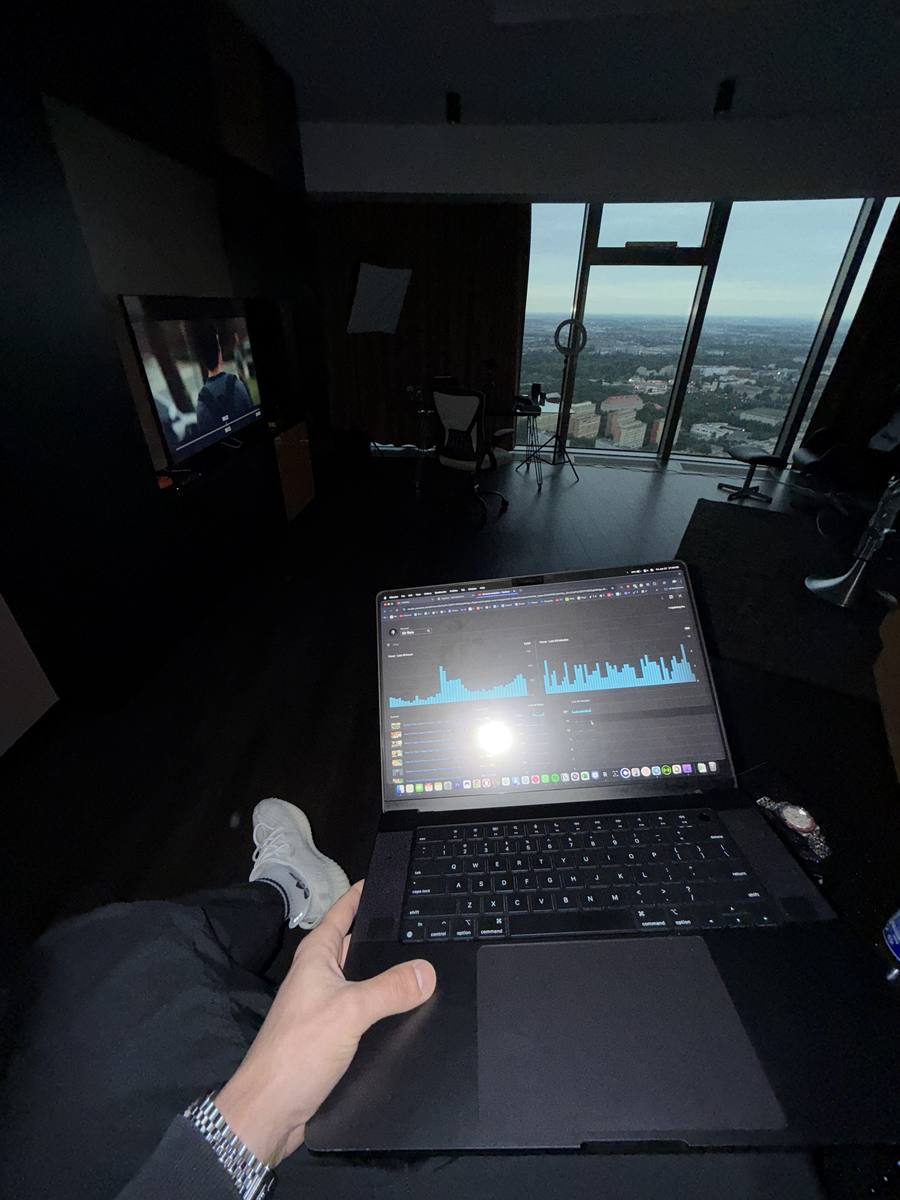 A dark studio at dusk with a city view through tall windows, a laptop showing analytics on Philipe's lap